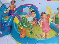 Photo showing children playing in the inflatable Dinoland water play center featuring a blue dinosaur arch sprayer, colorful balls, and splash zones.