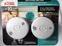 Photo of two white round Kidde smoke and carbon monoxide alarms in product packaging, highlighting 10-year battery life and talking/vocal feature.
