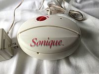 Top view photo of the Sonique hair remover main device showing oval shape, off-white plastic casing and red lettering.
