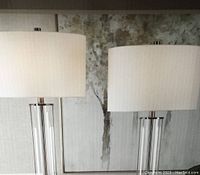 Pair of two new table lamps with clear glass cylindrical bases and white fabric drum shades placed in front of a painting.