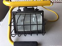 Yellow portable work flood light with metal protective cage and lens with textured glass, mounted on a frame with handle and yellow cable.