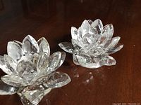 Two transparent glass flower candle holders with multiple faceted petals arranged in a blooming design on a dark wooden surface.