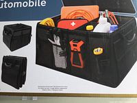Main product image showing the black rectangular automotive organizer with removable lid, various outer pockets with small items (gloves, spray bottle, flashlight) displayed inside for illustration only, and easy grip handles.