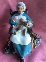Front view of Royal Doulton nanny figurine seated with teddy bear in lap against pink fabric background.