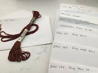 Closeup of single floss skein in brown color labeled 'DMC 300 Red Brown' and several white envelopes with handwritten labels of floss colors and DMC numbers.