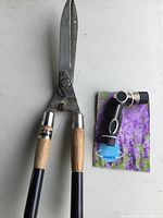 Garden shears with wooden handles and metal blades, showing signs of use, alongside new Orbit garden hose nozzle on purple floral packaging.
