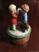 Side view of music box showing boy, girl, and dog figurine details on top of grassy base and wood-tone bottom.