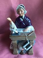 Front view of Royal Doulton porcelain figurine featuring elderly woman schoolmarm seated at desk with quill, paper, and objects underneath desk