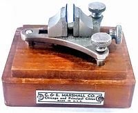 Close-up of the vintage watch movement holder mounted on the wooden base showing adjustable clamp and screws with company label on front.