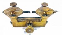 Front angled view of the vintage watch movement holder showing two triangular clamps with pointed prongs and knurled adjustment screws on a brass base.