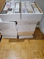 Photo showing 14 white cardboard storage boxes filled with baseball trading cards, stacked in two stacks on a wooden floor.