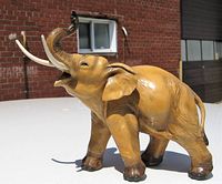 Side view of a brown porcelain wild elephant statue with trunk raised, showing detailed skin texture and white tusks, positioned outdoors with brick background.
