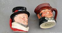 Side view of two tiny Toby jugs, Old Charley on the left with a black top hat and red coat, Beefeater on the right with dark red hat and black coat.