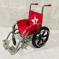 Side and front views of the American Girl doll wheelchair showing red fabric seat with white star, metal frame, missing front wheel, large black rear wheels, footrests and handles