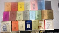 Photo showing 18 vintage glassware guide booklets arranged in two rows on a white surface. Various colors including orange, yellow, pink, blue, green, and tan with titles referencing pattern glass, carnival glass, and silver plate patterns.