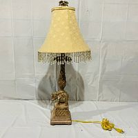 Full view of the elephant base lamp with beige, diamond-patterned fabric shade and hanging beaded fringe, lamp powered by yellow cord.