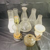 View of two oil lamps including one with electrical cord and several different shaped glass lamp shades in background.