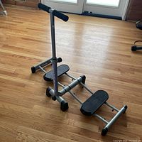 Angled side view of Leg Magic with handlebar, two foot pedals, and metal frame on wood floor.