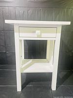 Front view of a white painted wooden side table with a drawer and lower shelf, photographed against a black backdrop.