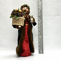 Full view of the figurine standing next to a ruler showing a height of approximately 12 inches. The male figure wears red and green robes with brown fur trim, holding a basket of fruit and a silver horn.