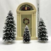 Full scene of decorated Christmas door with wreath and three snow-flocked Christmas trees of varying sizes.