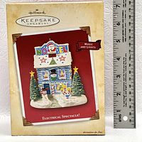 Photo of the original box for the Hallmark Keepsake ornament titled 'Electrical Spectacle' showing the product image, features music and lights, with a measuring ruler next to box.