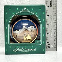 Photo showing Hallmark green box with clear circular window displaying the ornament; ornament has a nativity scene and camels, packaging is labeled 'Lighted Ornament Nativity' and measures approx 4.5 inches tall.
