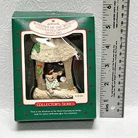 Photo of the 1987 Hallmark Windows of the World handcrafted ornament in original green box with clear plastic window, showing a violin playing figure under a thatched roof hut.