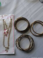 Photo showing six gold tone bangles with black and white accents arranged on a white cloth surface with a packaged gold tone chain necklace with floral packaging.