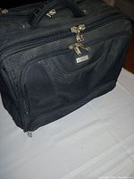 Front and side view showing black briefcase with diamond-patterned fabric and multiple compartments with zippers.