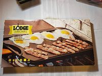 Front view of Lodge cast iron griddle box showing branding, product image of bacon and eggs cooking, and product dimensions 20x10 inches.