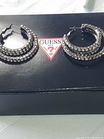 Pair of large Guess triple hoop earrings displayed on a black Guess box, showing front view with three rows of embedded clear crystals.
