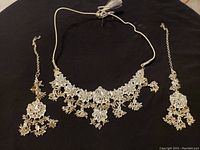 Full view of necklace centered with matching earrings on either side, showing silver-tone metal and clear rhinestone floral patterns with dangling bead clusters on black background.