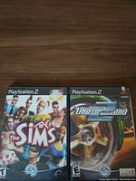 Two PlayStation 2 game cases for The Sims and Need For Speed Underground 2 viewed from the front on wooden surface.