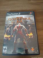 Closed view of God of War PS2 game case front cover with main character Kratos seen from behind holding chained weapons standing before ancient structures with flames.