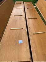 Two long oak wood planks side by side on outdoor grass surface, each with four narrow wooden slats across their width. The wood shows weathering and some surface damage.