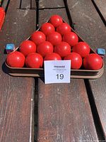 Fifteen red billiard balls arranged in a triangular metal rack tray on a wooden surface with two blue chalk cubes on each side.