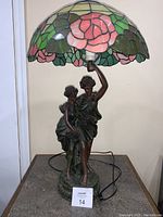 Full view of the decorative statue lamp with stained glass shade, showing the base statue of two women and the lamp cord.