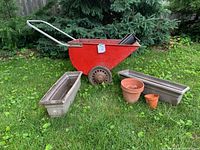 Red vintage garden cart with one wheel tire needing replacement, shown with two rectangular metal planter boxes and two small terracotta pots arranged on grass in front of pine trees.