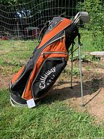 Photo showing the full Callaway golf bag standing on grass with clubs inside. Orange and black color scheme with zip pockets.