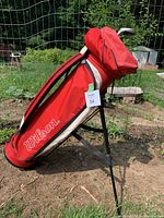 Red Wilson stand golf bag with leg supports set outdoors on dirt ground with green plants in background.