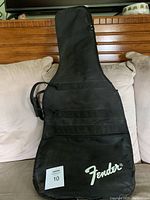 Soft sided black guitar case with Fender branding, standing upright on a couch with a wood-paneled background.