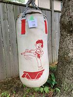 Off-white children's heavy bag featuring red cartoon of a karate child kicking, hung by metal chain on tree outdoors, showing signs of weathering and use.