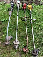Photo showing four gas-powered lawn trimmers with various handle styles laid on grass, showing overall length, wear, and cutting heads.