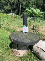 Photo of a heavy metal patio umbrella stand with a floral embossed design on the base and a black cylindrical holder pole in the center. The stand is placed outdoors on grass with garden fencing in the background.