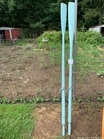Two light blue wooden rowing oars standing vertically outside, with metal oar locks attached and visible wear on paint.