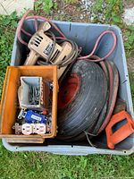 Container with power saw, red garden hose reel, large black wheel, yellow box with screws and electrical outlets, red-orange handle, and assorted hardware items seen outdoors on grass.