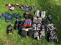 Top-down view of assorted hockey gloves, elbow pads, and shin guards arranged on grass. Various brands and colors visible with signs of wear.