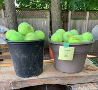 Photo of two small plastic containers filled with used Wilson US Open tennis balls placed on a wooden surface outdoors.