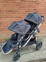 Side view of the Twin City Select baby jogger stroller showing two seats with black fabric and adjustable canopies on a sturdy aluminum frame with three wheels.
